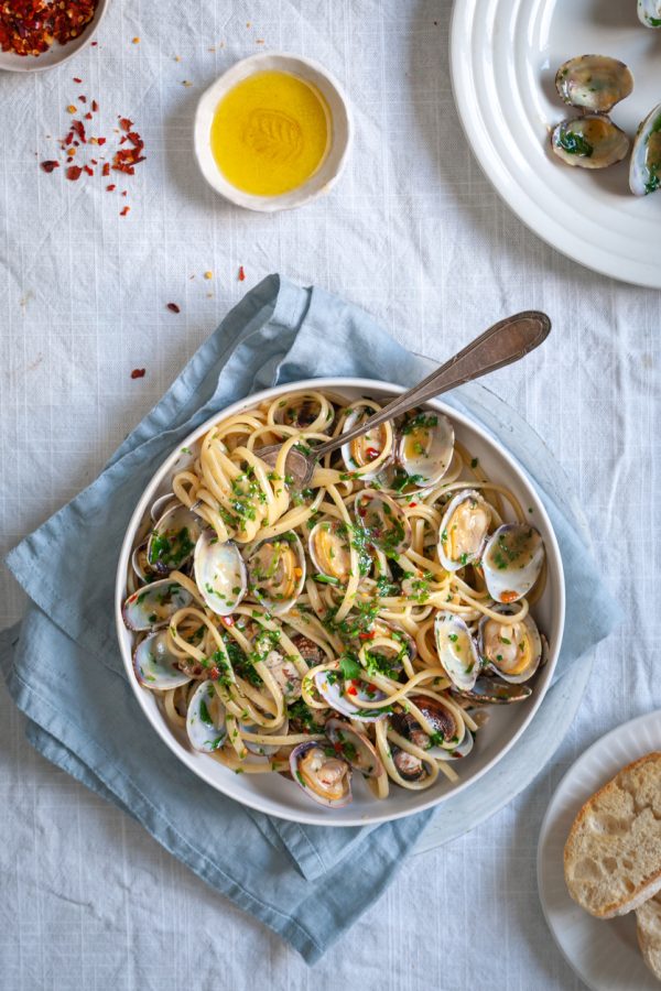 A bowl of pasta vongole on white table cloth with red chili pepper and olive oil